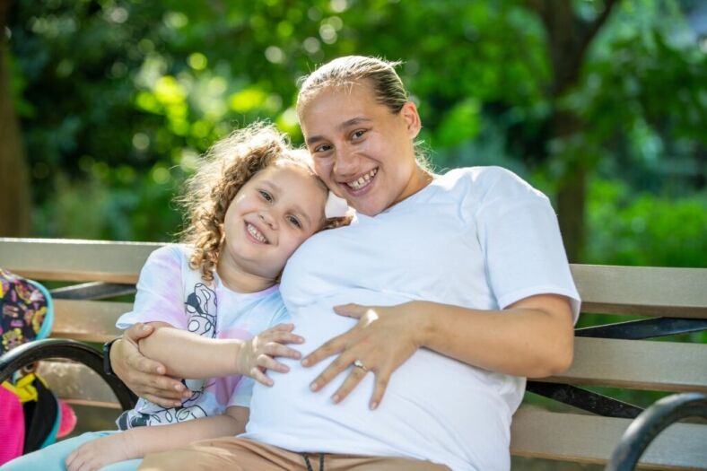 A pregnant survivor poses on a park bench with her young daughter.