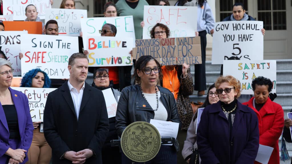 A woman, who is Safe Horizon’s Chief Program Officer Kelly Coyne, stands at a podium speaks to a crowd urging Governor Hochul to sign the SAFE Shelter Act into law. A group of people stands on steps behind her holding signs advocating for safety and access to shelter.