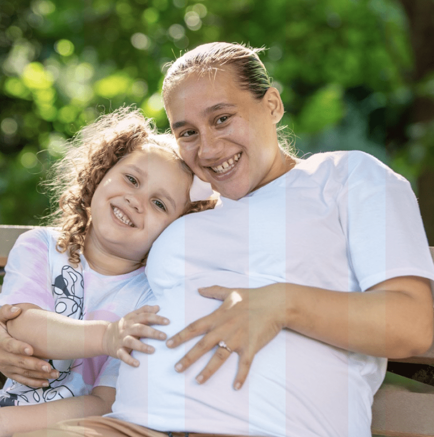 Pregnant woman and small girl smiling at the camera, with hands on pregnant belly.