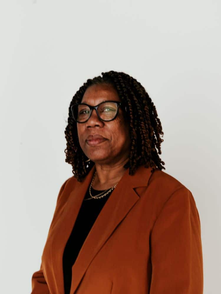 A woman with dark wavy hair wearing glasses and a brown blazer.