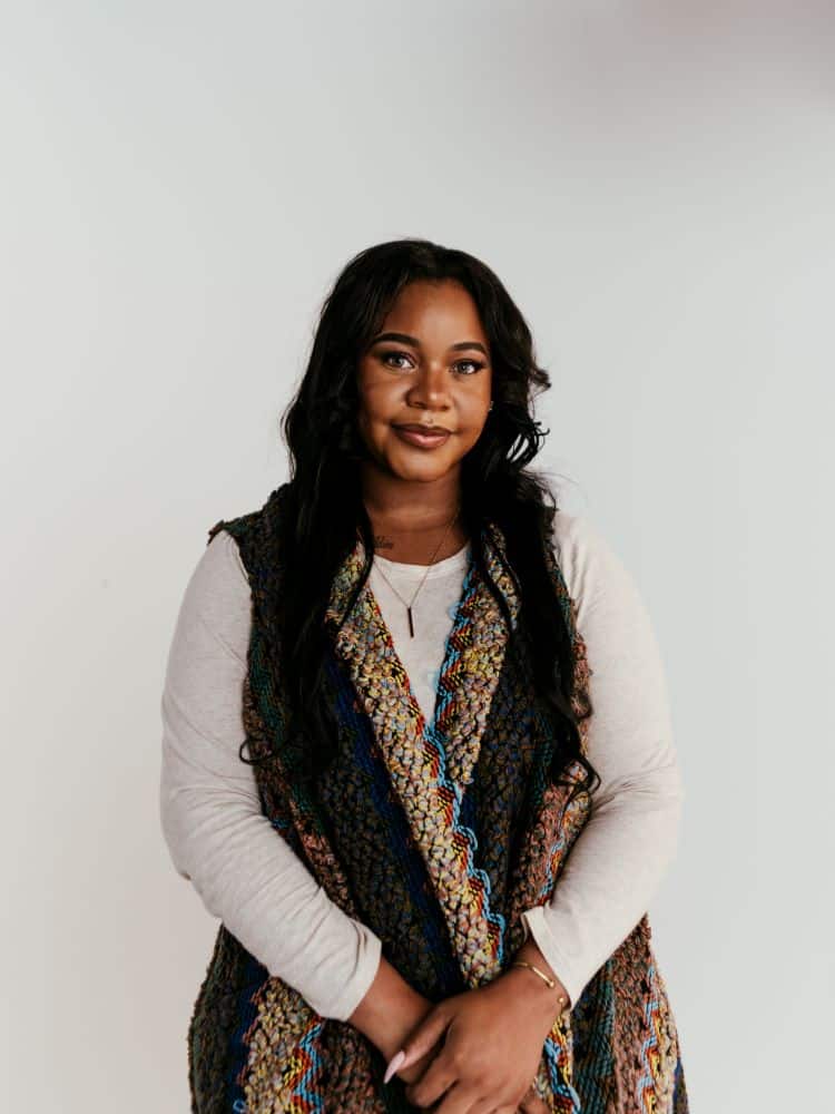 A woman with dark wavy hair wearing a white sweater and a brown vest.