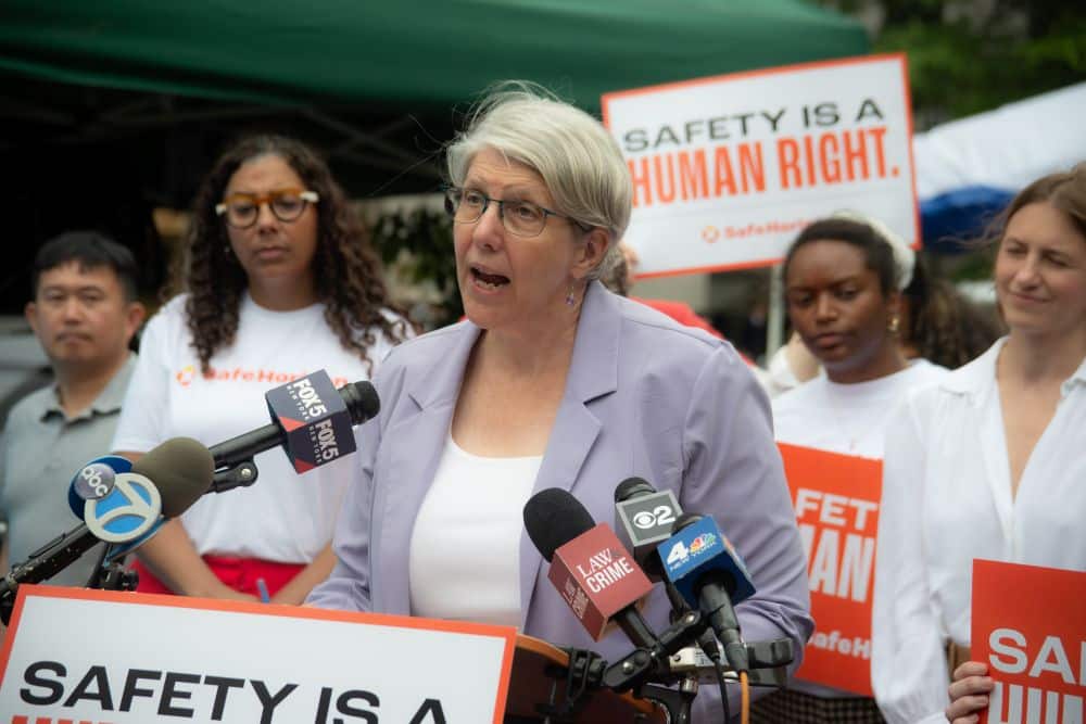 A woman wearing a lavender blazer speaks into a microphone behind a podium at a press conference. Signs reading 