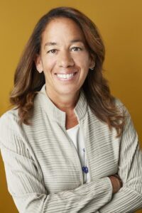 Headshot of a smiling woman with long brown hair, wearing a beige jacket.