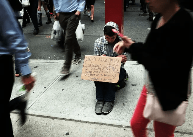 Young person sitting on the sidewalk holding a sign asking for help.
